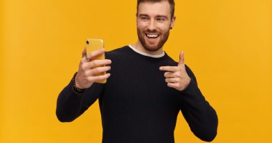 handsome-guy-with-brunette-hair-beard-has-piercing-wearing-black-sweater-making-selfie-has-video-call-pointing-finger-screen-watching-his-phone-isolated-yellow-wall