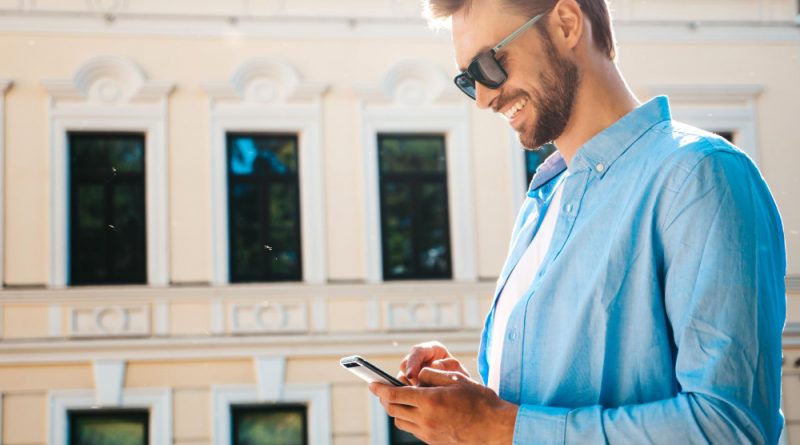 Handsome Smiling Stylish Hipster Lambersexual Modelmodern Man Dressed Blue Shirt Fashion Male Posing Street Background Outdoors Sunset Using Smartphone Apps Sunglasses