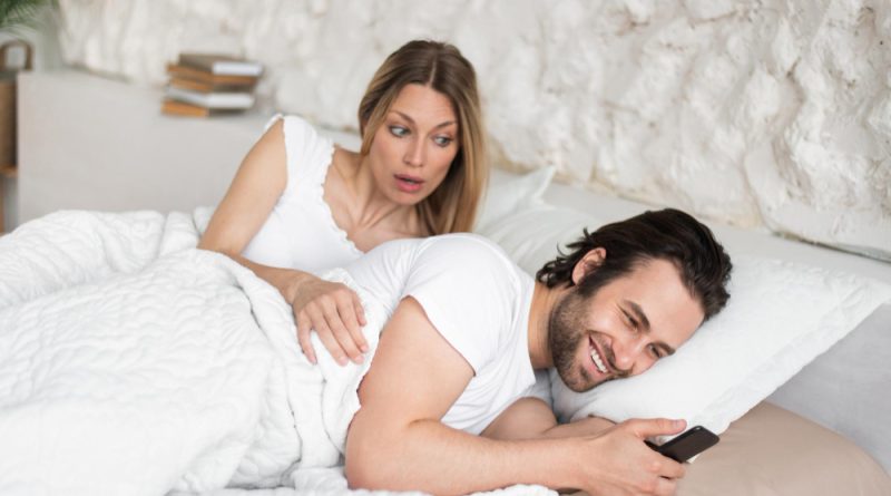 Shocked young woman seeing her husband messaging his lover smartphone while lying bed