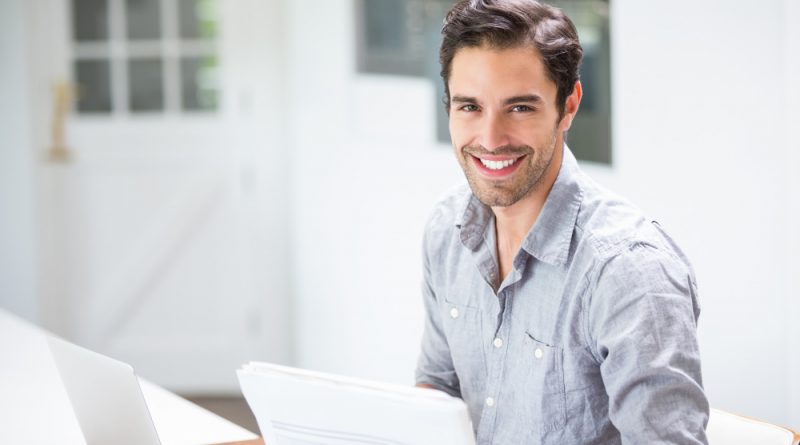 Smiling Young Man Holding Documents While Sitting Desk With Laptop